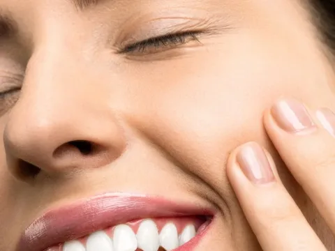 A close-up portrait of a joyful woman with closed eyes, pink lipstick, and her hand touching her cheek.