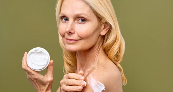 An image of a mature woman smiling while applying moisturizing cream on her shoulder embodies a healthy skincare routine and antiaging care for radiant skin